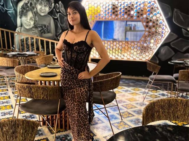 AshlyLee stands in a funky cafe wearing a black lace top and leopard print pants, hands on her hips, with wavy black hair and a confident look.
