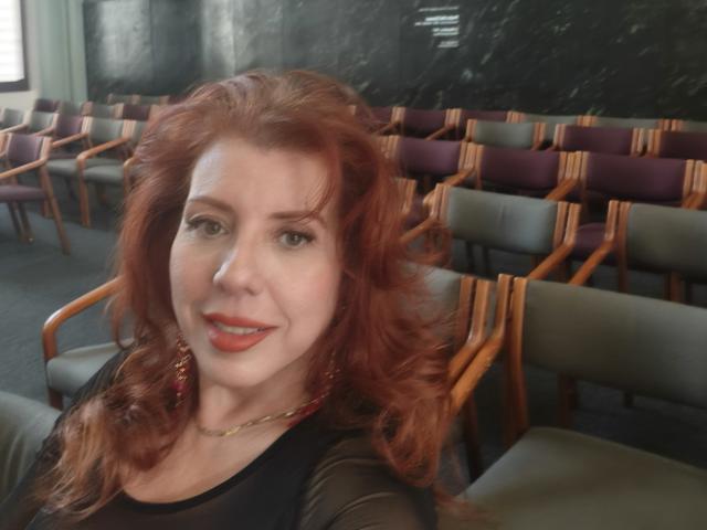 Khharha with red hair, smiling, wearing a black sheer top, red earrings, and a gold necklace, sitting in a room with wooden chairs and purple cushions.