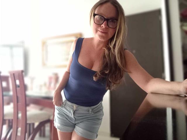 PamelaColin with long blonde hair, glasses, blue tank top showing her tits, and light blue shorts, leaning forward, smiling, hands in pockets, standing in a room.