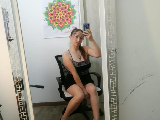 GianineLarose in a black dress, sitting on a chair, taking a mirror selfie with her phone, showing her tits and pussy, in a room with a mandala poster.