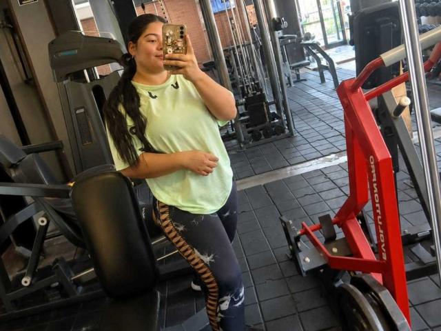 ElizaWiliamst with long dark hair, green shirt, black and patterned pants, standing in gym, holding phone, mirror selfie, red exercise bike, black tiled floor, serious expression.