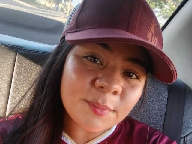 ElizaWiliamst with brown eyes, straight black hair, wearing a maroon baseball cap, smiling slightly, has light brown skin, and is in a car with grey leather seats.