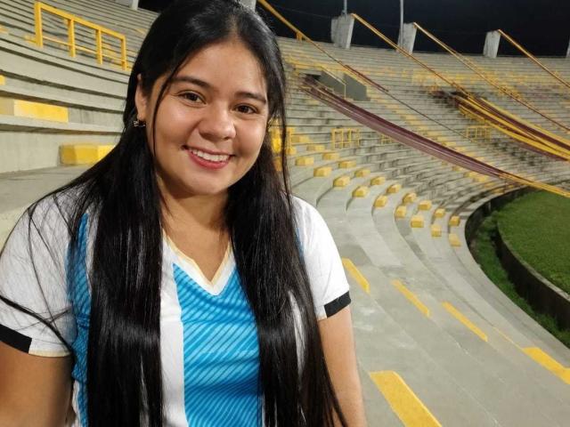ElizaWiliamst has long black hair, a cute smile, and is wearing a blue and white sports shirt. Her tits are small and she's standing in a stadium.