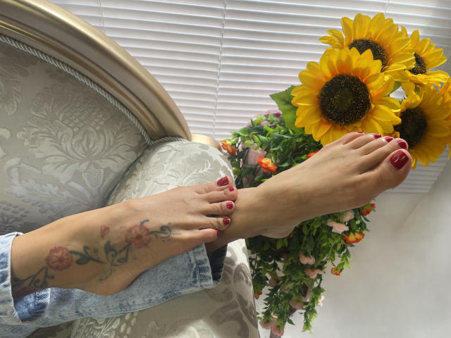 Os pés de MiaAmoree, com unhas pintadas de vermelho e uma tatuagem floral no tornozelo, descansam em uma cadeira branca, com girassóis ao fundo..