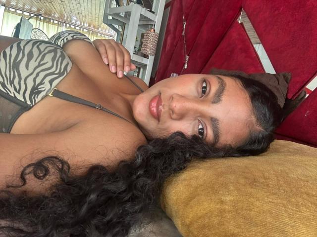 NatalyBlonder with curly black hair, brown eyes, and tan skin, lying on a yellow pillow, wearing a zebra-patterned top, showing her small tits and a cute nose.