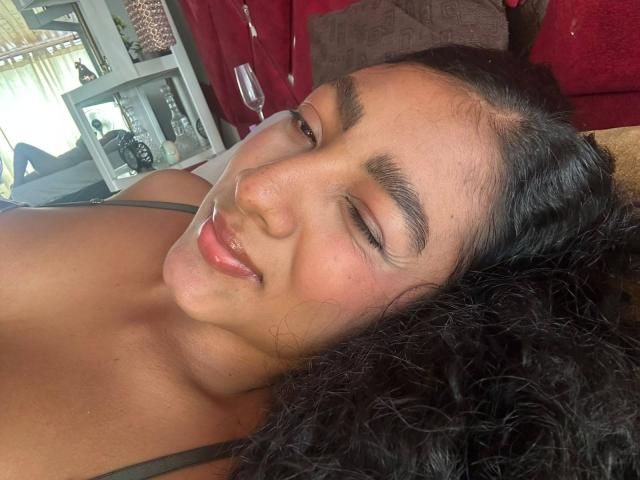 NatalyBlonder's face, winking with a slight smile, has curly black hair, medium brown skin, and is wearing a gray tank top, lying down with a red background.
