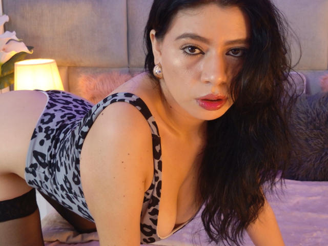 LindaMancini with wavy black hair, wearing a leopard-print lingerie top, on all fours, showing her ass, in a bedroom with a lit lamp in the background.