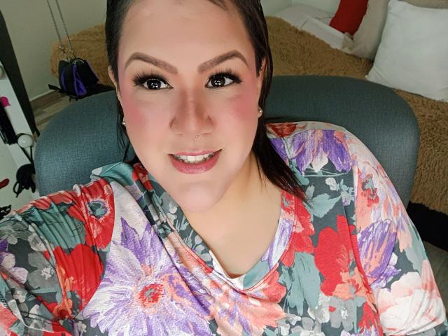 GiiannaMorrys with dark brown eyes, pink blush, and floral shirt, smiling at camera. She has a nose ring and long brown hair. Her tits are partially visible.