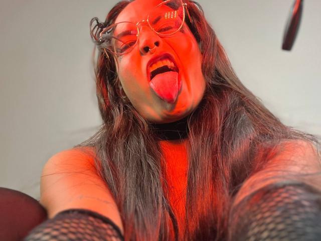 LilithRussoo with red-lit face, tongue out, wearing glasses, and fishnet sleeves, has long brown hair and a nose ring, taking a selfie. Boobs and ass are partially visible.
