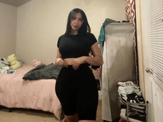 QueenLatinBeauty, avec des cheveux noirs, des lunettes, et un haut noir court ainsi que un pantalon noir taille haute, se tient dans une chambre avec un lit rose et des jouets en peluche..