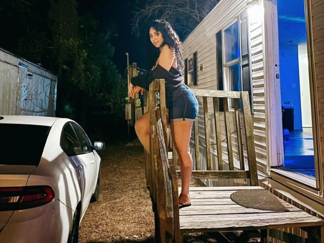 Une femme, QueenLatinBeauty, avec des cheveux noirs bouclés, un haut noir à une épaule et un short en jean, debout sur des marches en bois, regardant en arrière, à côté d'une voiture blanche, la nuit, dans une remorque éclairée..