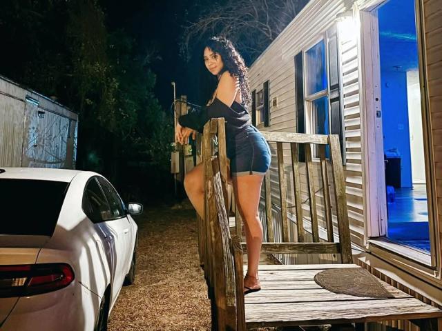 QueenLatinBeauty, avec ses cheveux noirs bouclés, portant un haut noir à une épaule et un short en jean, se tient sur un porche en bois, appuyée sur la rambarde, à côté d'une voiture blanche, la nuit..