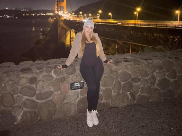 QueenLatinBeauty, avec de longs cheveux roux, portant un bonnet blanc, une veste beige, un body noir, des leggings noirs et des baskets blanches, est appuyée contre un mur de pierre la nuit..