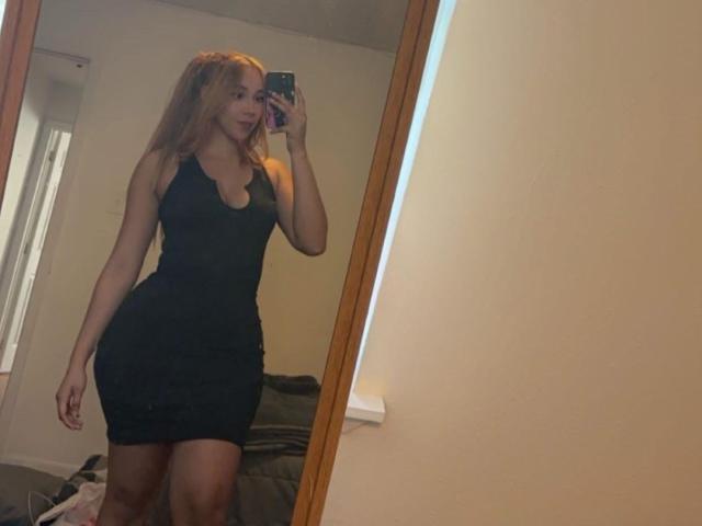 QueenLatinBeauty with long blonde hair, wearing a tight black dress, showing off her big ass and small tits in a mirror selfie, holding a phone.