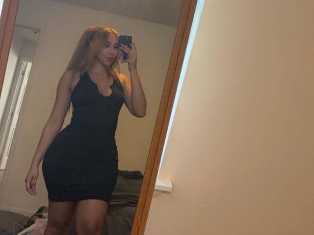 QueenLatinBeauty, avec de longs cheveux roux, portant une robe noire moulante, montrant ses courbes et sa poitrine généreuse, prenant un selfie devant un miroir avec un smartphone dans une chambre..