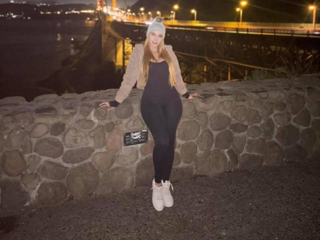 QueenLatinBeauty avec de longs cheveux blonds, un bonnet beige, une veste marron, un body noir, des leggings noirs et des bottes blanches, appuyée contre un mur de pierre la nuit, les bras reposant, souriante, debout..