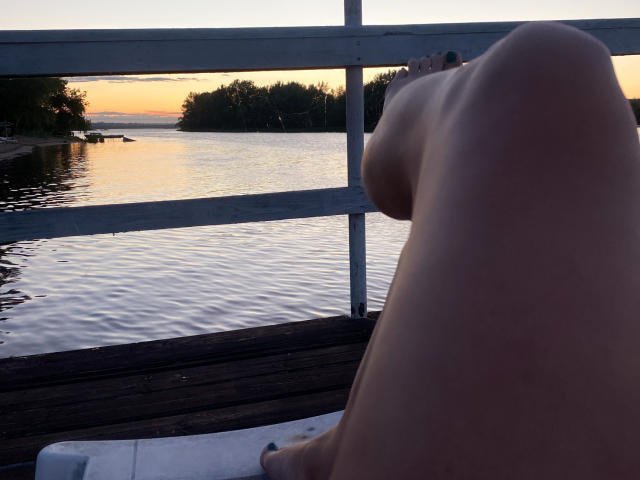 EvelynCutes nackte Beine und Gesäß im Vordergrund, mit sichtbaren Zehen, sitzend auf einem hölzernen Steg bei Sonnenuntergang, mit Blick auf ruhiges Wasser im Hintergrund..
