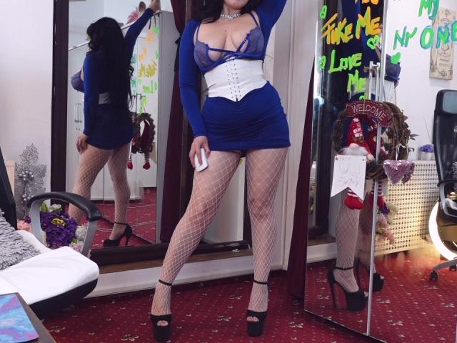 LexyRene stands in a room, wearing a blue sheer dress with white corset, fishnet stockings, and black high heels. Her boobs are visible through the dress. Her ass is prominent.
