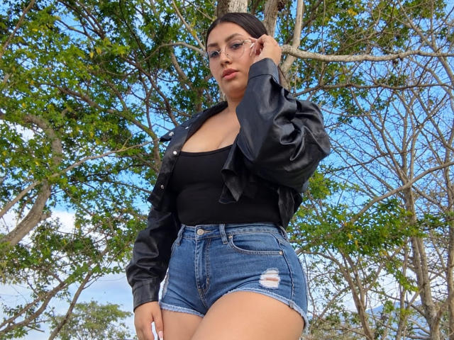 Catalina Rivero mit Brille, schwarzem Tanktop, schwarzer Jacke und Jeansshorts, steht neben einem Baum, eine Hand im Haar, blauer Himmel und grüne Blätter im Hintergrund, wobei ihre Brüste sichtbar sind..