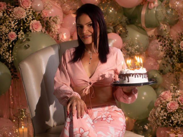 KatteRouse, con cabello oscuro, un top corto rosa y pijamas florales rosas, sentada en una silla, sosteniendo un pastel con velas, sonriendo, con globos rosas y rosas en el fondo..