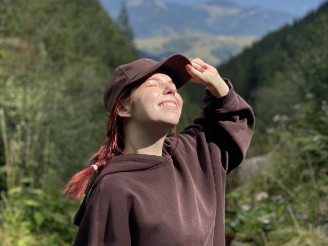 AlexXas with red braids, smiling, wearing a brown cap and hoodie, raising her cap to her head, showing her teeth, in a sunny, green, forested landscape.