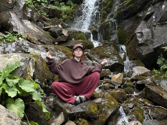 AlexXas, sitting cross-legged on mossy rocks, brown hoodie, red pants, hands in meditation pose, waterfall behind, smiling, green foliage on left, wet rocks, overcast.