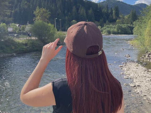 AlexXas with red hair, wearing a brown baseball cap and black shirt, standing by a rocky river, holding her cap with her right hand.