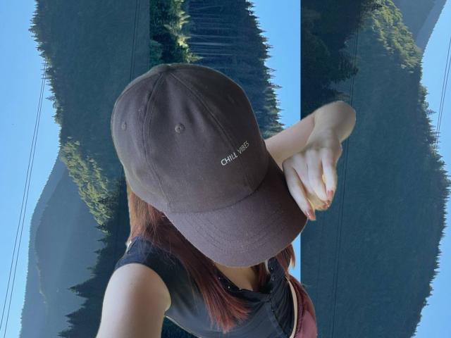 AlexXas wearing a brown cap, black shirt, and red nails, with arms crossed, reflected in a glass window showing green trees and a clear blue sky.