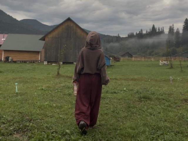 AlexXas, back to camera, in brown hoodie and maroon pants, walking on green grass towards a wooden barn, holding a bottle in right hand, overcast sky, mist in background.