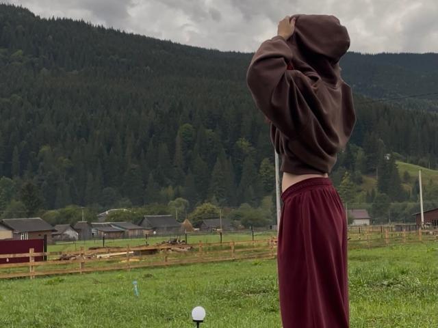 AlexXas, met een bruine hoodie, rode broek en een ontbloot buikje, staat in een groen veld, met de rug naar de camera gericht, handen in de capuchon, bewolkte lucht, beboste heuvels, houten omheining op de achtergrond..