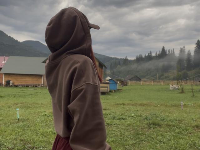 AlexXas stands with back to camera, brown hoodie, cap, red skirt, grassy field, cloudy sky, mountains, wooden houses, and misty forest in the background.