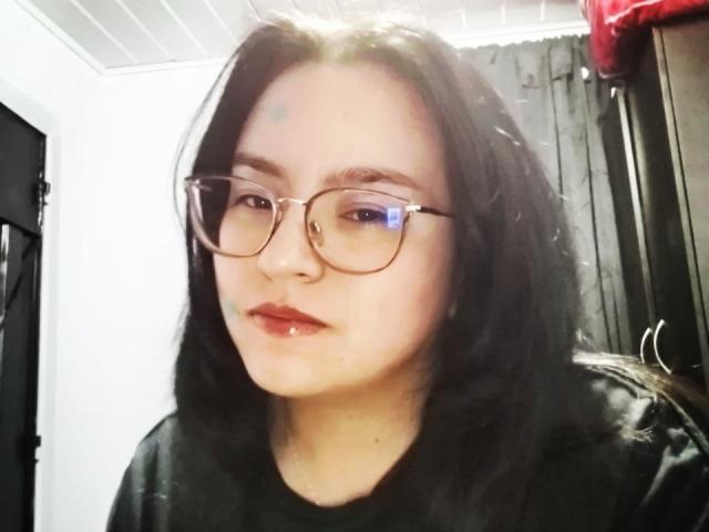 AmberColombo with glasses, black hair, pale skin, small blue mark on forehead, black shirt, red lips, slight drool on chin, ceiling light reflecting off glasses.