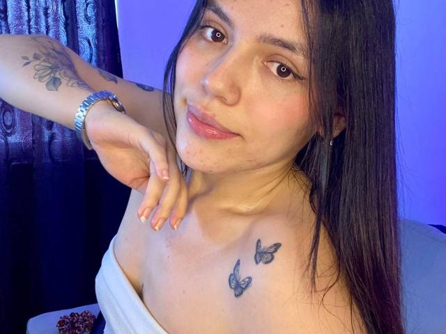 SophiaMarx has a cute face with long dark hair, small nose ring, subtle smile, and butterfly tattoos on her chest. She's wearing a white off-shoulder top, showing her tits.