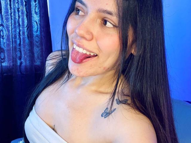 SophiaMarx with long black hair, nose ring, and tattoo on right shoulder, sticking out tongue, wearing white strapless top, blue background. Boobs partially covered by top.