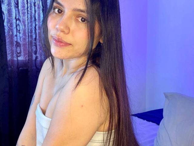 SophiaMarx has long brown hair, fair skin, and small tits, wearing a white strapless top, smiling slightly with a slight acne mark on her cheek, in a blue-lit room.