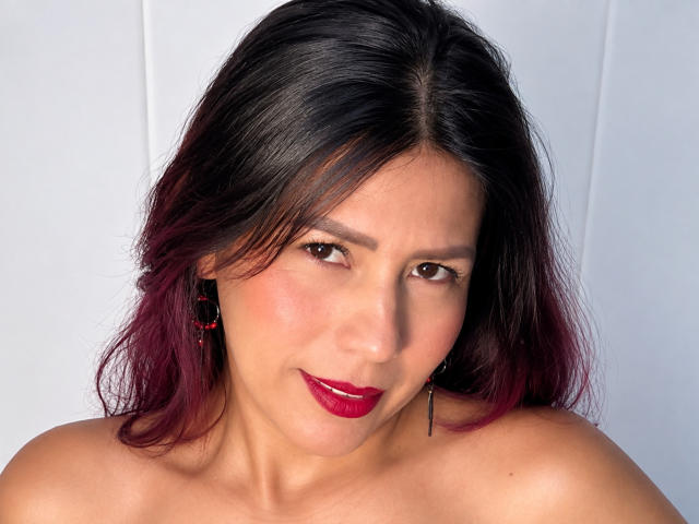 LunaTomson has dark brown eyes, red lipstick, and shoulder-length black hair with red tips. She's looking at the camera with a slight smile, wearing red earrings.