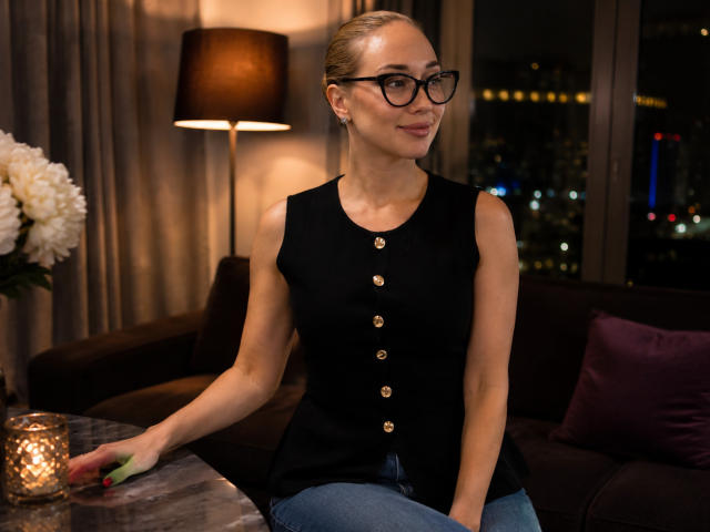 AngelicElite, blonde, wearing black sleeveless top with gold buttons, blue jeans, black glasses, sitting on couch, hand on table, lamp and city lights in background, smiling.