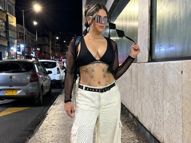 RedMoont, avec des tatouages sur son ventre, un soutien-gorge noir laissant entrevoir ses seins, un haut transparent à manches longues, un pantalon blanc à pois, une ceinture noire et des lunettes de soleil, tenant un miroir, debout dans une rue la nuit..