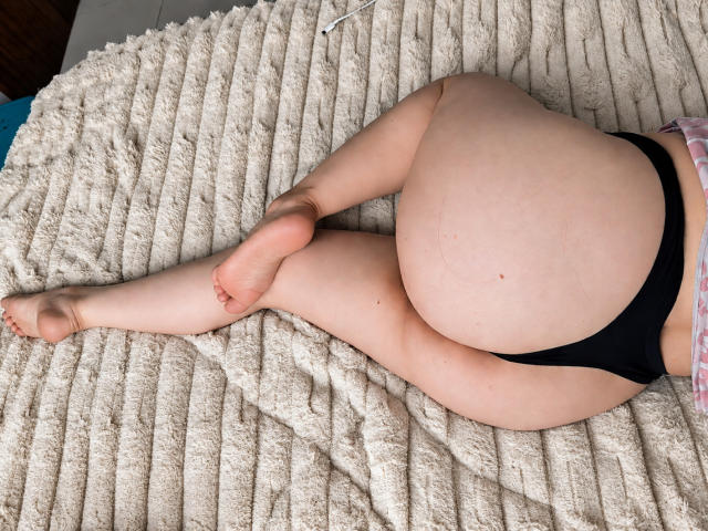 BellaFournierHot lies on a beige, ribbed blanket, wearing black panties, with her ass and legs visible, her feet crossed at the ankles, showing pale skin and a small mole.