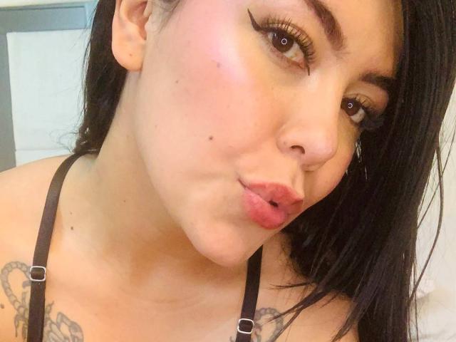 CatalinaReiyna with pouty lips, black hair, and tattoos, wearing a black strap top, showing her tits and small mole on her left cheek, in a selfie.