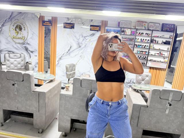 Elena Saenz, avec ses cheveux roses, son débardeur de sport noir et son jean bleu, exhibe ses abdominaux tonifiés et ses petites seins, en prenant un selfie devant un miroir dans un salon avec des chaises grises..