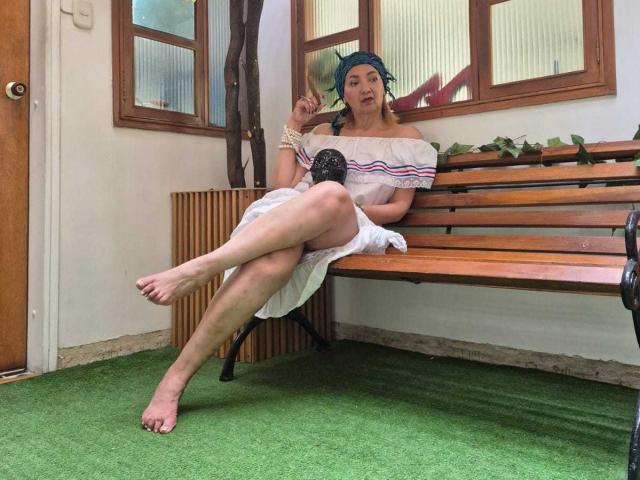 AmelieLux with bare feet, white off-shoulder dress, blue headscarf, holding black mask, legs crossed, sitting on wooden bench, green carpet, wooden door, windows, and plant in background.