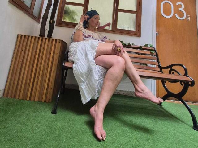 AmelieLux sits barefoot on a wooden bench, wearing a striped dress and headscarf, with legs crossed, showing her ass and hairy legs, in a relaxed, homey setting.