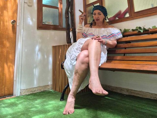 AmelieLux, barefoot, sitting on wooden bench, wearing white dress with red and blue stripes, holding cigarette, green headscarf, dirty leg, green grass, wooden door, windows, relaxed, smoking.