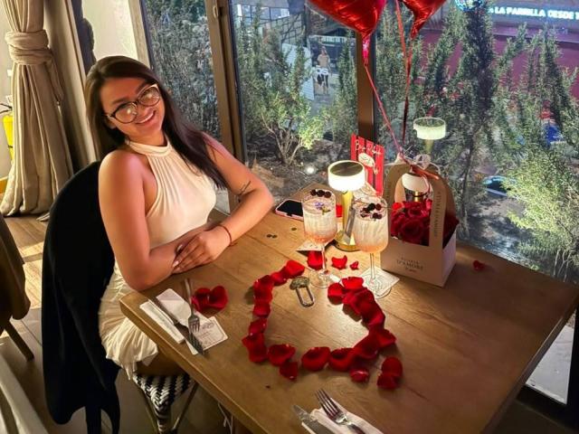 AlessiaFerry, con gli occhiali, indossa un abito bianco senza maniche, seduta a un tavolo di legno con petali di rosa, dolci al cioccolato e palloncini rossi, sorride alla fotocamera..