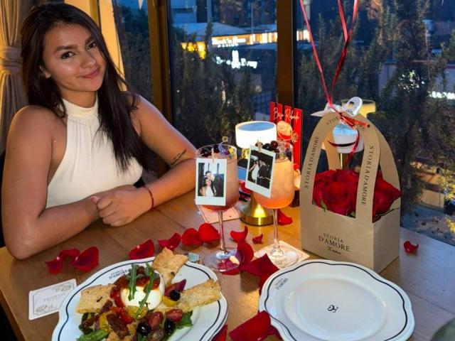 AlessiaFerry mit dunklem Haar, einem weißen Top ohne Ärmel, lächelnd an einem Tisch mit Speisen und Getränken, die mit Rosenblättern verziert sind, sowie einem Geschenkkorb mit roten Rosen und einem weißen Band..