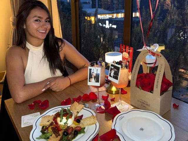 AlessiaFerry with tits up, smiling, wearing a white halter top, at a table with rose-petaled chips, photo frame, cocktail, and brown box with roses.
