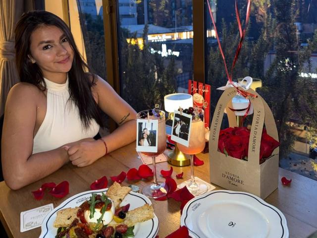 AlessiaFerry with tits and long dark hair, smiling, wearing a white sleeveless top, at a table with rose petals, flowers, and a photo on a glass.