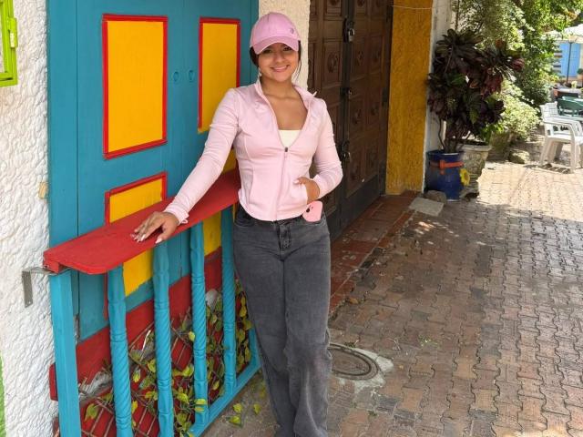 AlessiaFerry stands with a pink cap, pink zip-up, and gray pants, leaning on a colorful railing, smiling, with her hand on her belly, showing her tits.