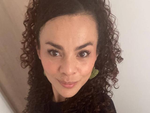 MissSunnyShine met krullend bruin haar, een medium huidskleur en groene oorbellen, lacht naar de camera en laat haar borsten subtiel zien met een schattige, zelfverzekerde uitdrukking..